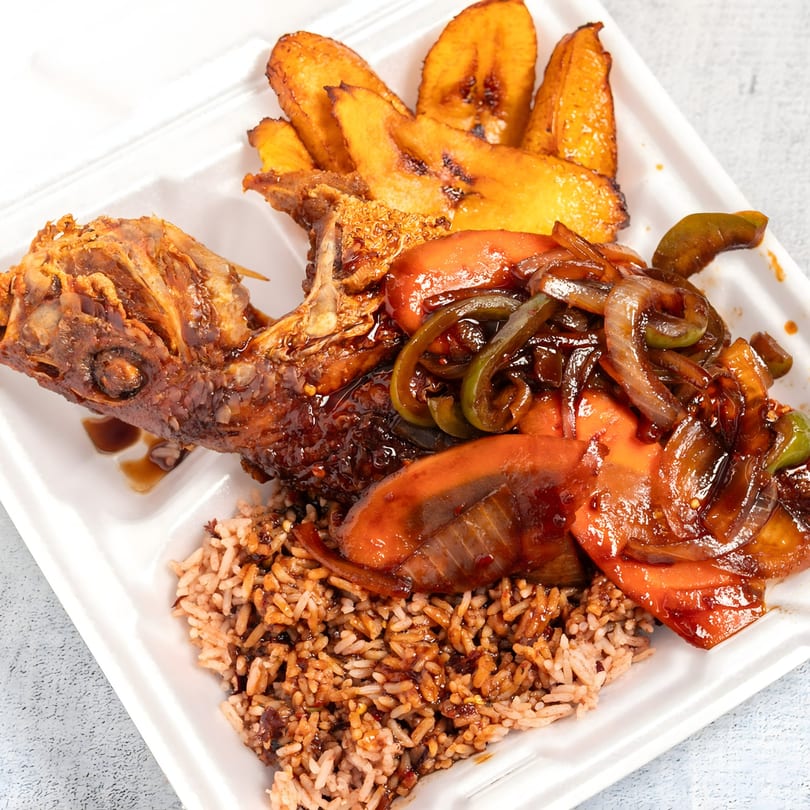 Jamaican escovitch fish with brown rice, fried plantains, and sautéed vegetables in a takeout container.