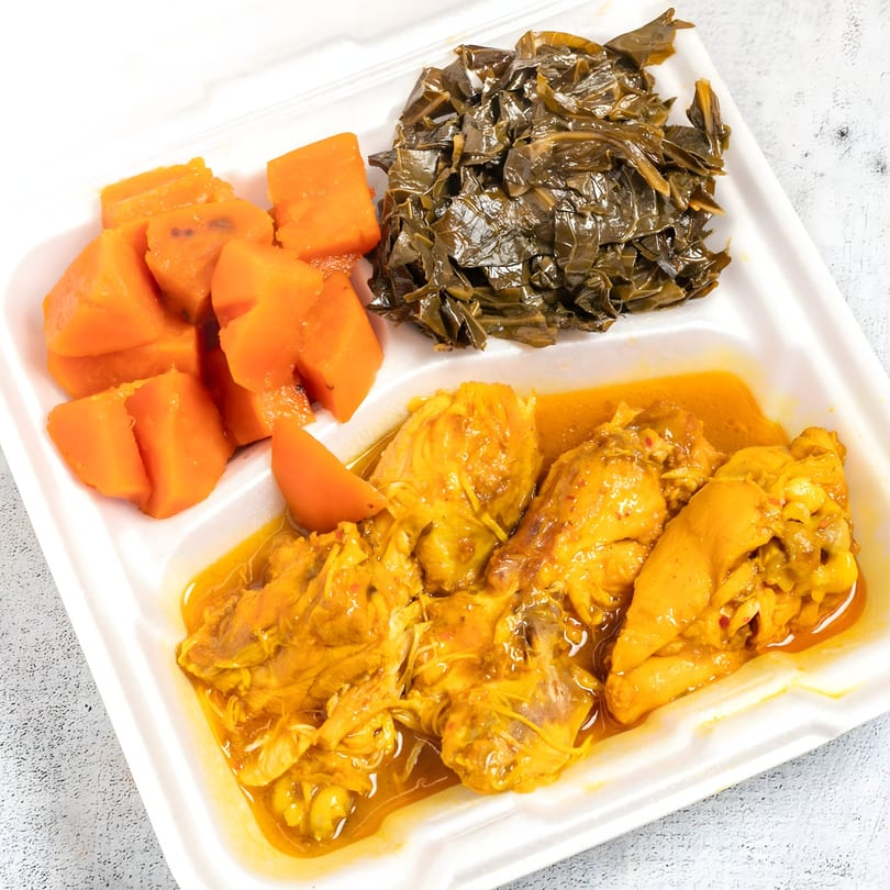 Curry chicken with collard greens and sweet potatoes in a white takeout container.
