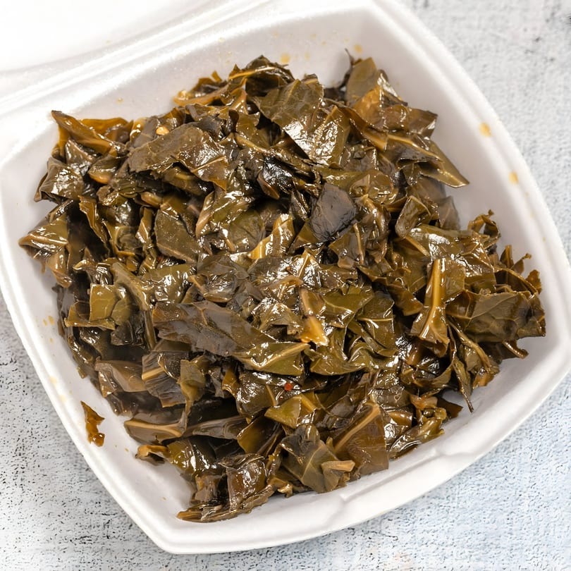 A white takeout container filled with a generous serving of glistening, dark green collard greens.